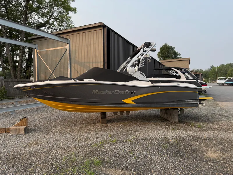 Slide: The Image of 2015 MasterCraft X10 boat with black cover, parked on gravel near storage buildings. - 0