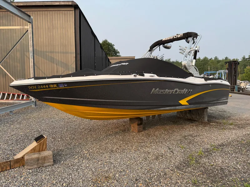 Slide: The Image of 2015 MasterCraft X10 boat with black and yellow design, parked on gravel. - 2