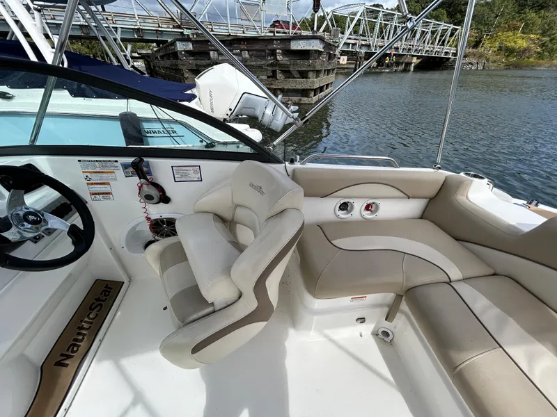 Slide: The Image of 2021 NauticStar 203DC Sport Deck interior with beige seating and steering wheel, docked by a bridge. - 18