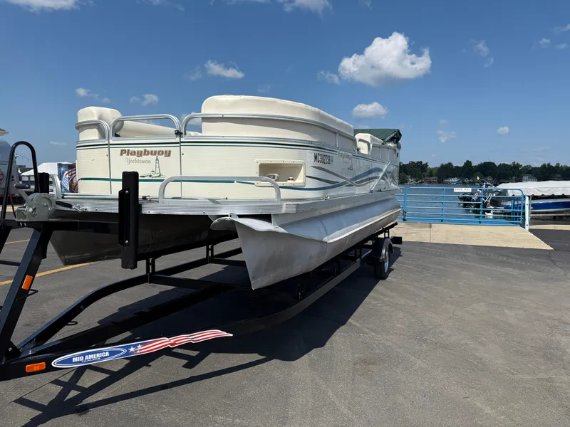Slide: The Image of 2000 Playbuoy Yachtsman 21 pontoon boat on trailer, parked near a marina under blue sky. - 3