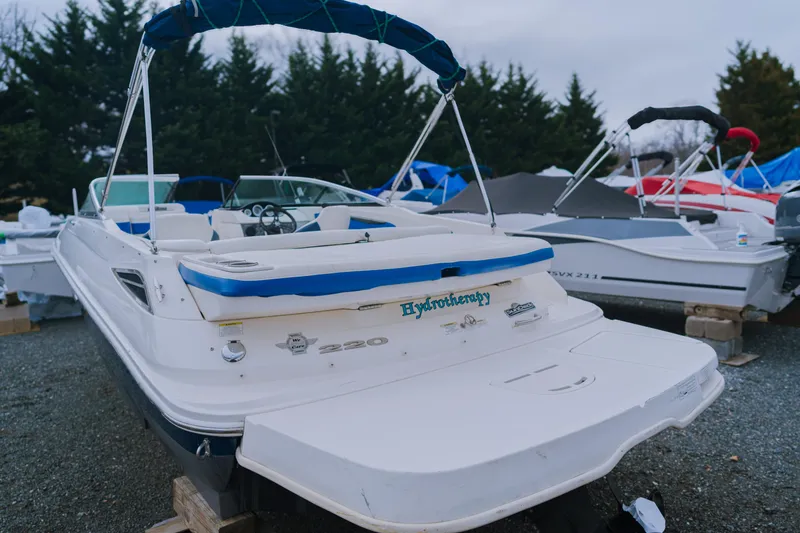 Slide: The Image of 2013 Sea Ray 220 Sundeck boat with blue accents, parked outdoors on gravel. - 2