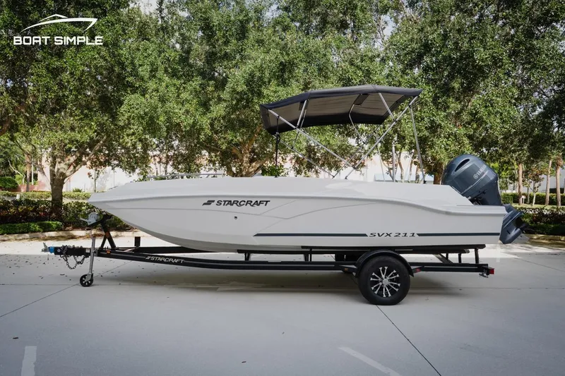 Slide: The Image of 2023 Starcraft SVX 211 OB boat on trailer, parked outdoors with trees in background. - 0