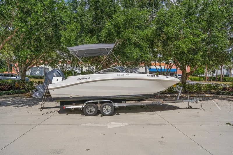 Slide: The Image of 2025 Hurricane SunDeck 217 OB boat on trailer, parked outdoors with trees in background. - 1