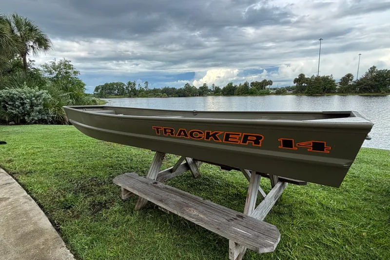 The Image of 2026 Tracker Grizzly 14 Jon boat displayed on a wooden stand by a scenic lake. - 0