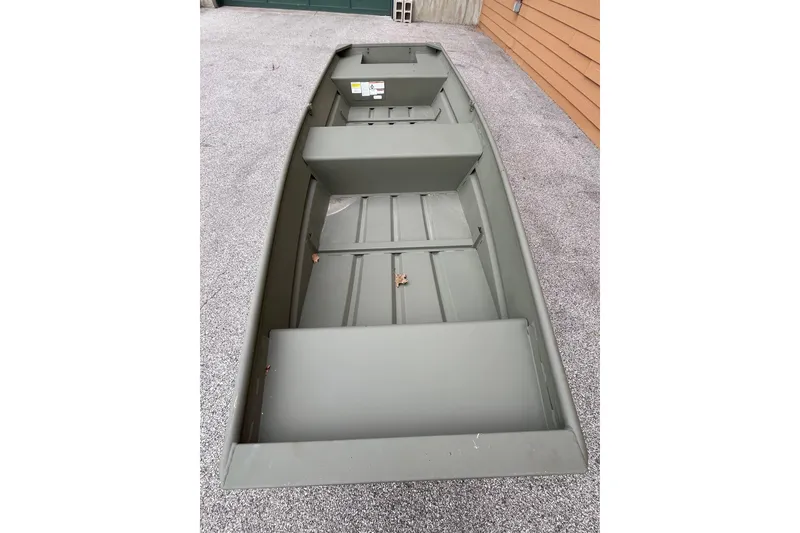 Slide: The Image of 2026 Tracker Grizzly 14 Jon boat, olive green, parked on gravel near wooden wall. - 2