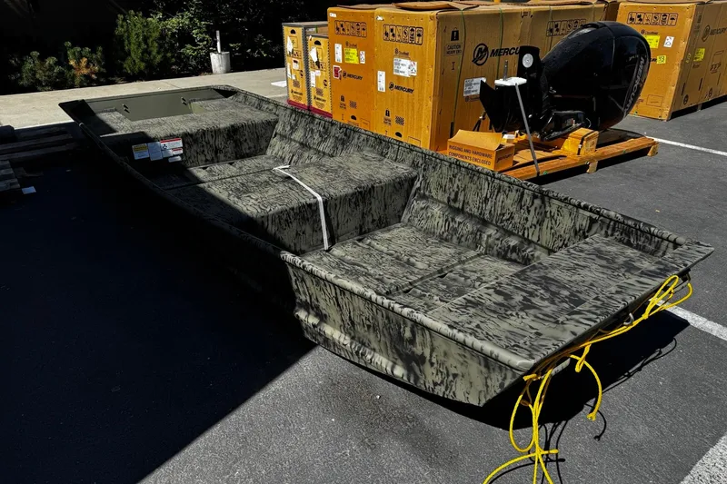 Slide: The Image of 2026 Tracker Grizzly 15 Jon boat in camouflage, parked outdoors with shipping crates nearby. - 2