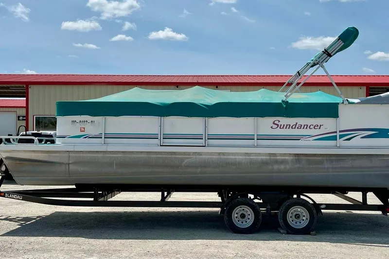 Slide: The Image of 1998 Sundancer Pontoons 240 boat with green canopy on trailer, parked outdoors. - 13