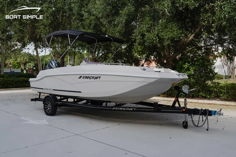 Slide: The Image of 2023 Starcraft SVX 211 OB boat on trailer, parked outdoors with trees in background. - 4