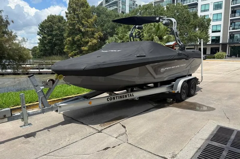Slide: The Image of 2018 Nautique Super Air Nautique GS22 boat on trailer, covered, near water and buildings. - 2