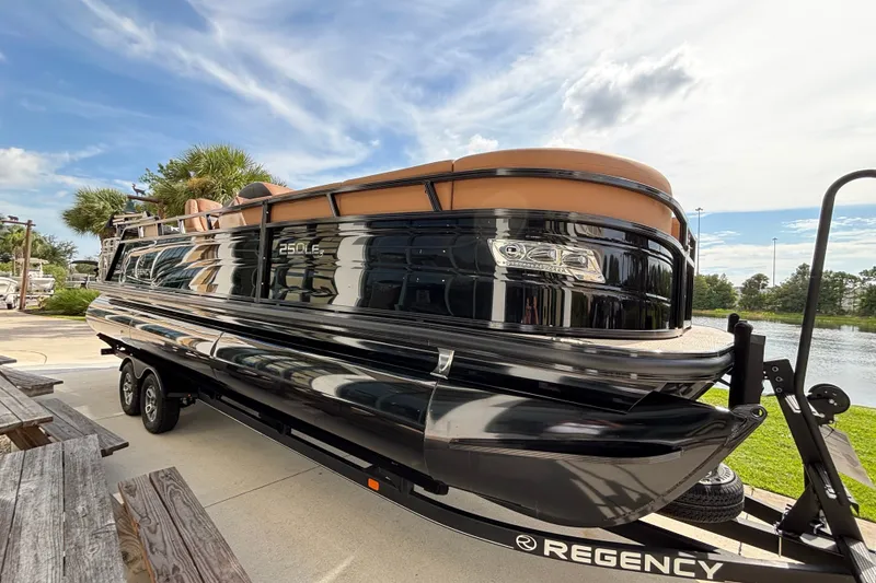 The Image of 2025 Regency 250 LE3 Sport pontoon boat on trailer, parked near a scenic waterfront. - 0