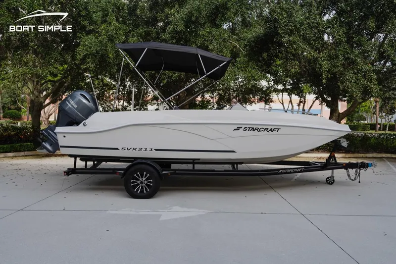 Slide: The Image of 2023 Starcraft SVX 211 OB boat on trailer, parked outdoors with trees in background. - 3