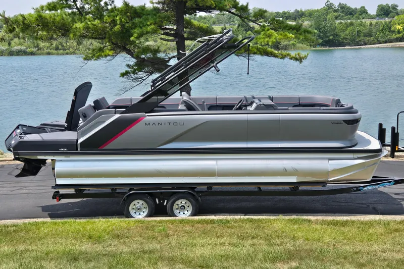 The Image of 2024 Manitou Explore pontoon boat on trailer by a scenic lake. - 0