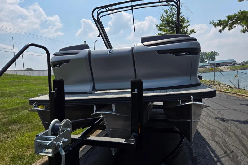 Slide: The Image of 2024 Manitou Explore pontoon boat on trailer, parked near a lake under a cloudy sky. - 6