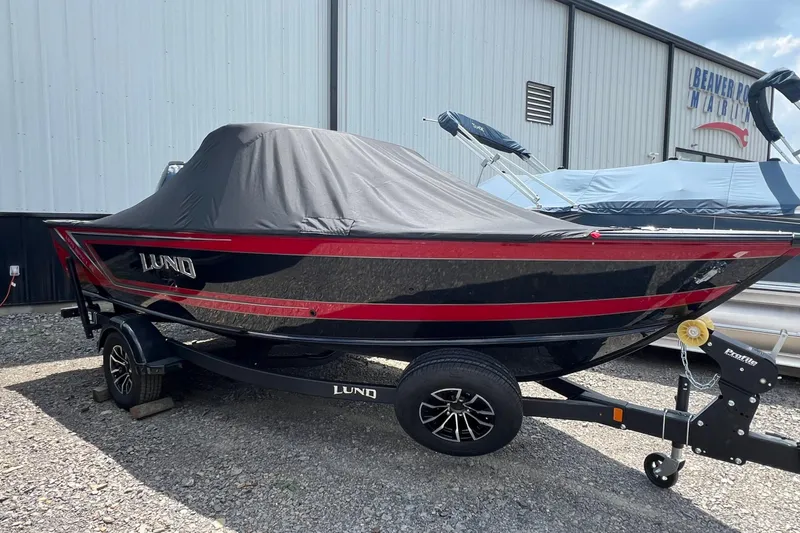The Image of 2026 Lund 1875 Impact XS Sport boat with cover, parked on a trailer outside a marine facility. - 1