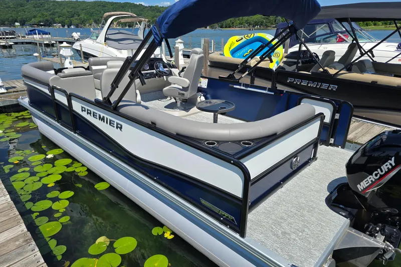 Slide: The Image of 2025 Premier 230 Sunsation RE pontoon boat docked on a lake with lush surroundings. - 6