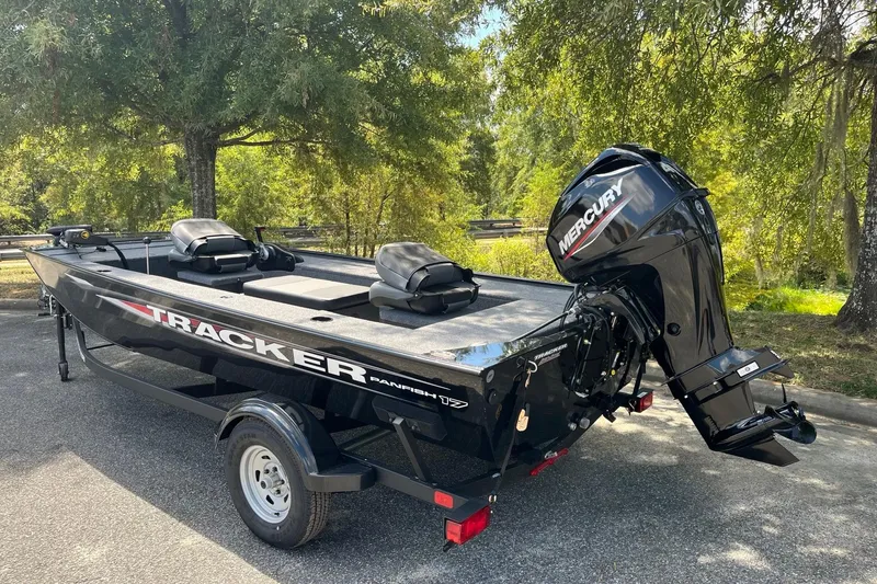 Slide: The Image of 2025 Tracker Panfish 17 boat with Mercury engine, parked outdoors under trees. - 2