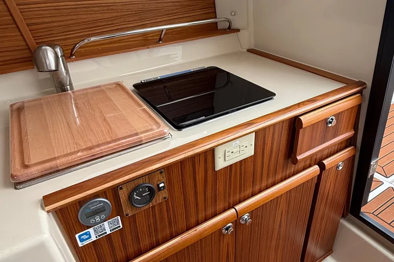 Slide: The Image of 2025 Ranger Tugs R-25 galley with wooden cabinetry, sink, and electric cooktop. - 33