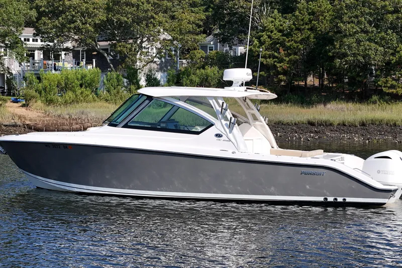 Slide: The Image of 2018 Pursuit DC 295 Dual Console boat on calm water near a wooded shoreline. - 2