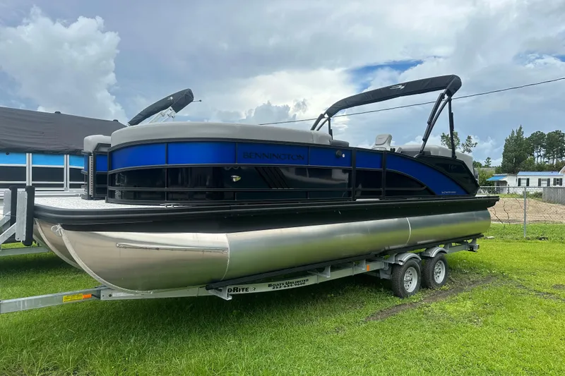Slide: The Image of 2025 Bennington 24M pontoon boat on trailer, parked on grass under cloudy sky. - 1