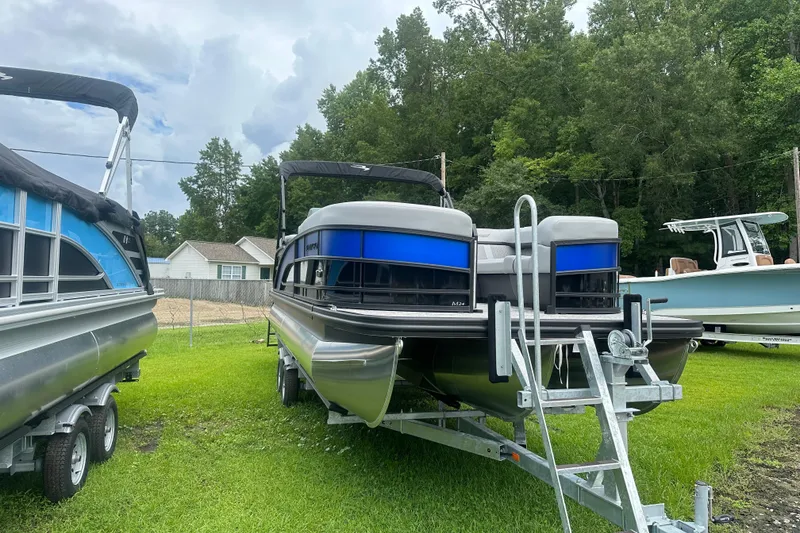 Slide: The Image of 2025 Bennington 24M pontoon boat on trailer, parked on grass with trees in background. - 2