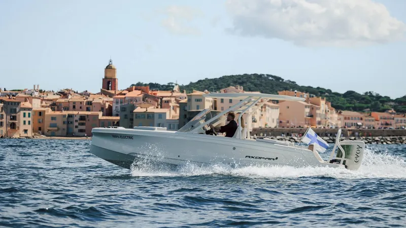 Slide: The Image of 2025 Axopar AX/E 22 T-Top boat cruising near coastal town, scenic waterfront view. - 0