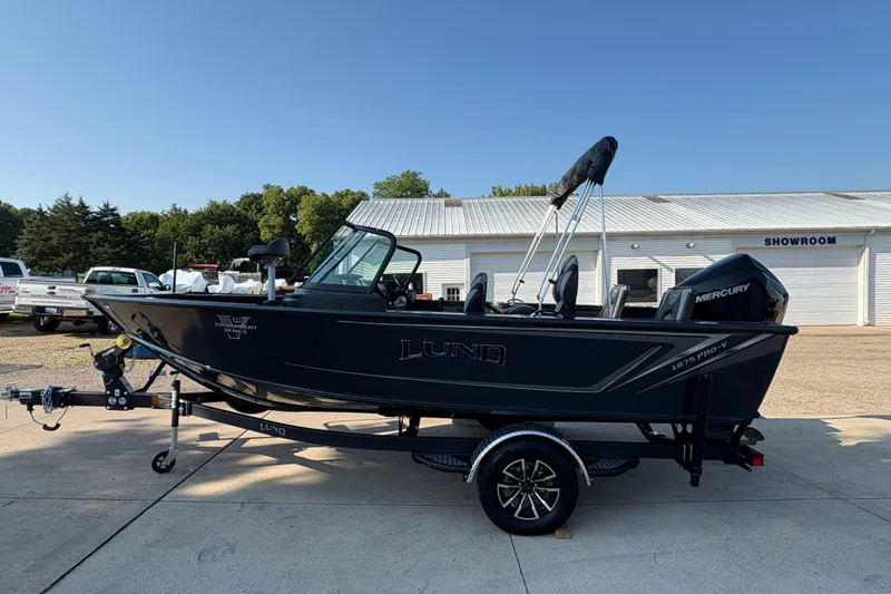 Slide: The Image of 2026 Lund 1875 Pro-V Sport boat on trailer outside showroom, featuring sleek black design. - 4