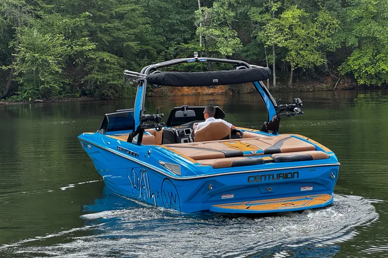 Slide: The Image of Blue 2025 Centurion Nv233 boat cruising on a calm lake surrounded by lush greenery. - 3