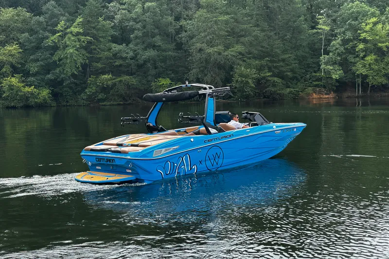 Slide: The Image of 2025 Centurion Nv233 boat cruising on a serene lake surrounded by lush trees. - 5