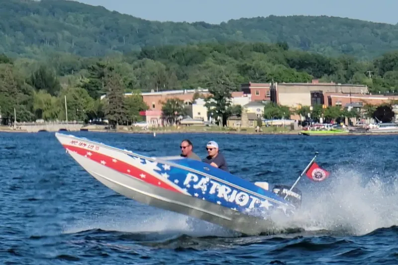 Slide: The Image of Speedboat "Patriot" on water, two people onboard, scenic town and hills in background. - 3