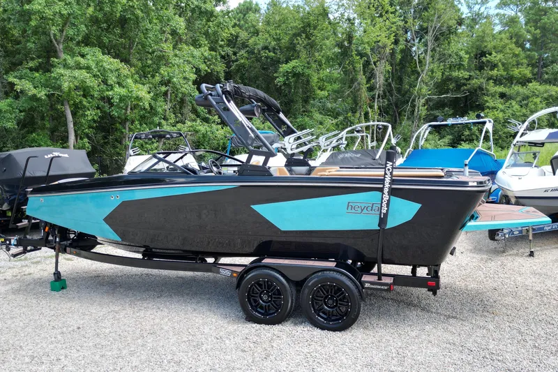 Slide: The Image of 2022 Heyday WTSurf boat in black and teal, parked on a gravel lot with trees in background. - 3