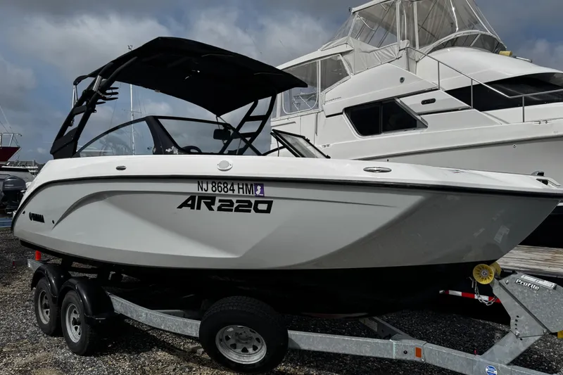 Slide: The Image of 2023 Yamaha Boats AR220 on trailer, parked near larger yacht, under cloudy sky. - 5