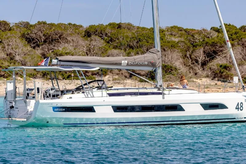 Slide: The Image of 2026 Dufour 48 yacht sailing in clear blue waters near a forested shoreline. - 6