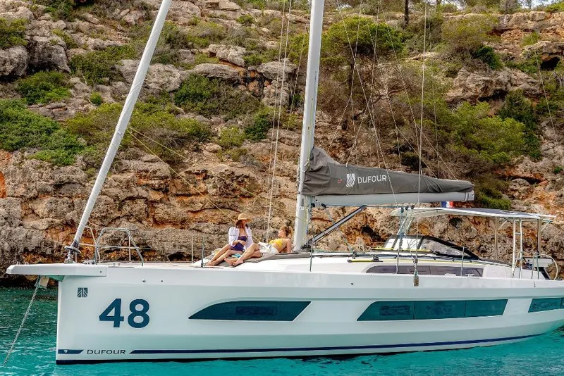 Slide: The Image of 2026 Dufour 48 yacht anchored near rocky coastline with lush greenery. - 7
