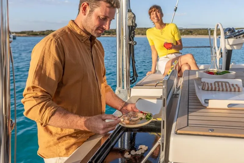 Slide: The Image of Man cooking on Dufour 48 yacht, woman enjoying drink, ocean backdrop, 2026 model. - 13