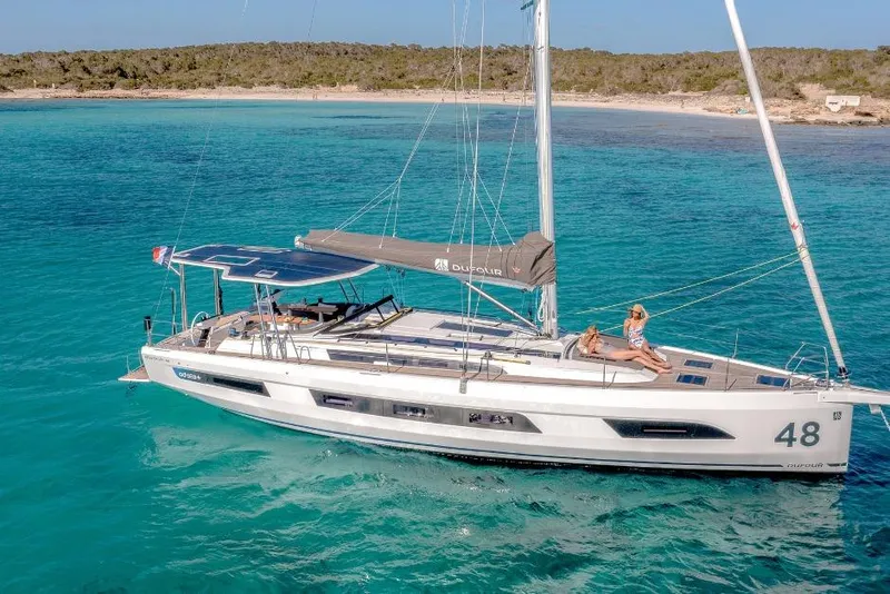 Slide: The Image of 2026 Dufour 48 yacht sailing in clear turquoise waters near a sandy beach. - 2