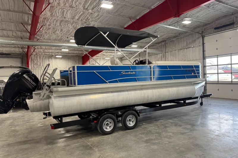 Slide: The Image of 2019 Sweetwater 2486 pontoon boat with blue panels, on a trailer in a spacious indoor facility. - 3