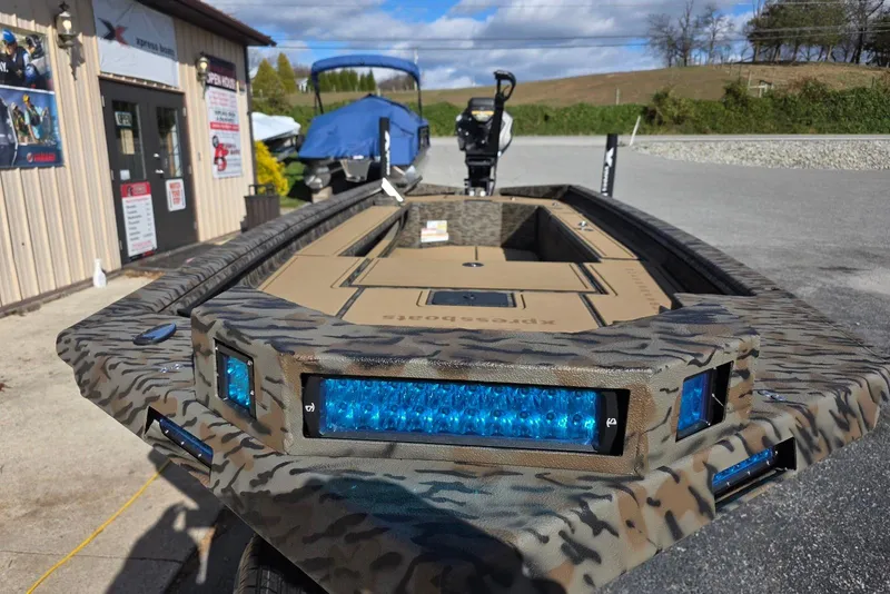 Slide: The Image of 2026 Xpress Bayou 180 boat with camouflage design and blue LED lights, parked outdoors. - 23