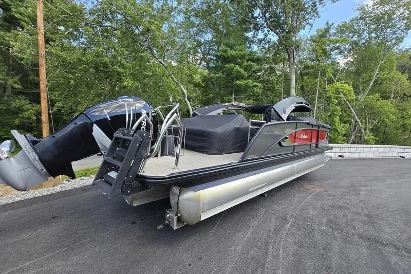 Slide: The Image of 2023 Barletta L25UCA pontoon boat parked on asphalt, surrounded by trees. - 1