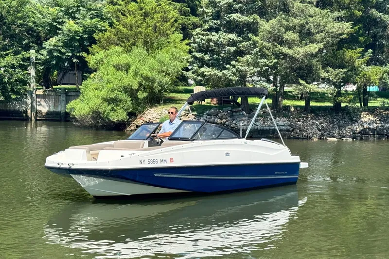 Slide: The Image of 2017 Bayliner 190 Deck Boat cruising on a calm river with lush greenery. - 0
