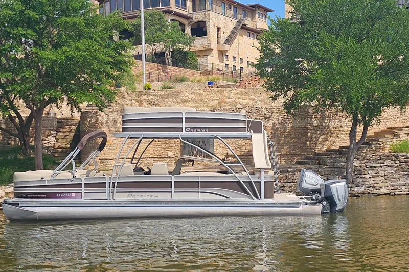 The Image of 2015 Premier 310 Escalante pontoon boat on a calm lake near a stone building. - 1