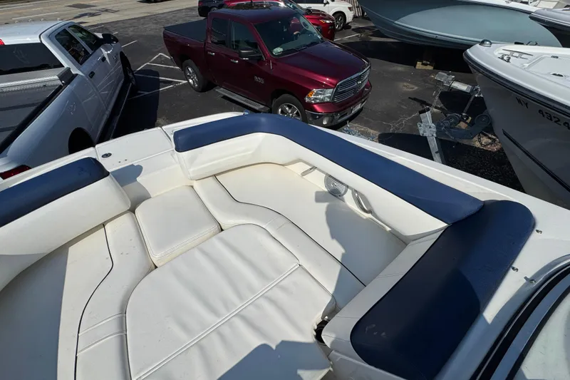 Slide: The Image of 2015 Bayliner 190 Deck Boat interior with white and blue seating, parked near trucks. - 19