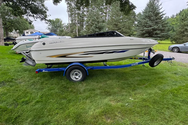 The Image of 2008 Glastron MX175 boat on a blue trailer, parked on grass with trees in the background. - 1