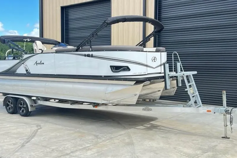 Slide: The Image of 2024 Avalon LSV 2585 pontoon boat on trailer, parked outside a building. - 1