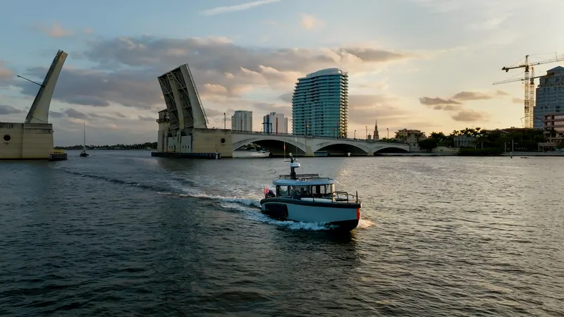 Slide: The Image of 2023 Arksen 30 boat passing under raised drawbridge at sunset, city skyline in background. - 22