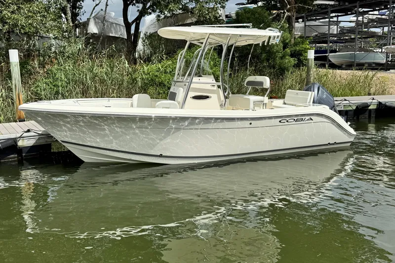 The Image of 2026 Cobia 220 Center Console boat docked on calm water, surrounded by greenery. - 0