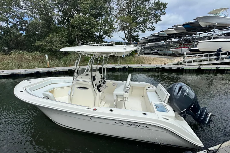 Slide: The Image of 2026 Cobia 220 Center Console boat docked in a marina, surrounded by trees and other boats. - 2
