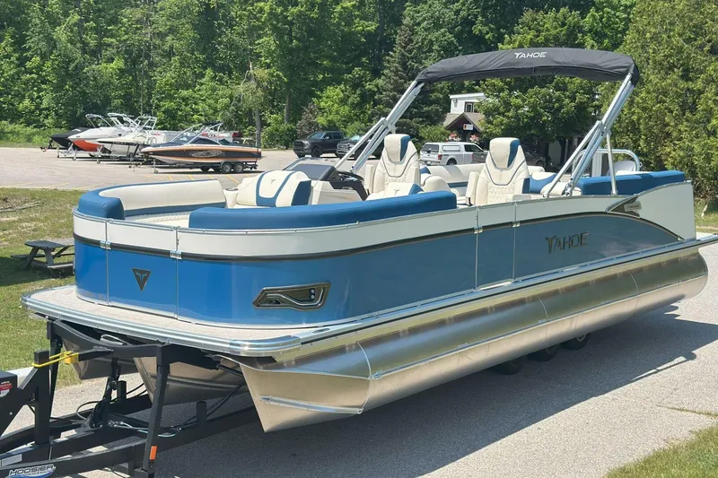 Slide: The Image of 2025 Tahoe 2585 pontoon boat with blue and white design on a trailer, surrounded by greenery. - 3