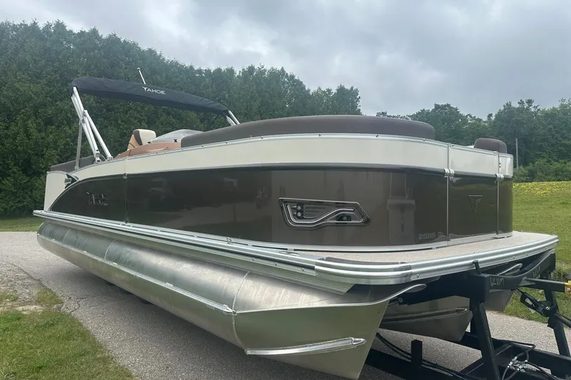 Slide: The Image of 2025 Tahoe 2585 pontoon boat on trailer, parked on a grassy area with trees in background. - 9