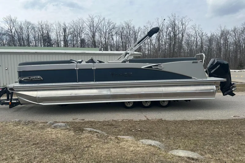 Slide: The Image of 2025 Tahoe 2585 pontoon boat with Mercury engine, parked on a trailer near a wooded area. - 0
