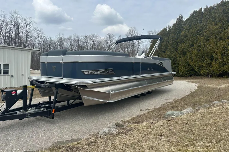 Slide: The Image of 2025 Tahoe 2585 pontoon boat on trailer, parked outdoors near trees and building. - 1
